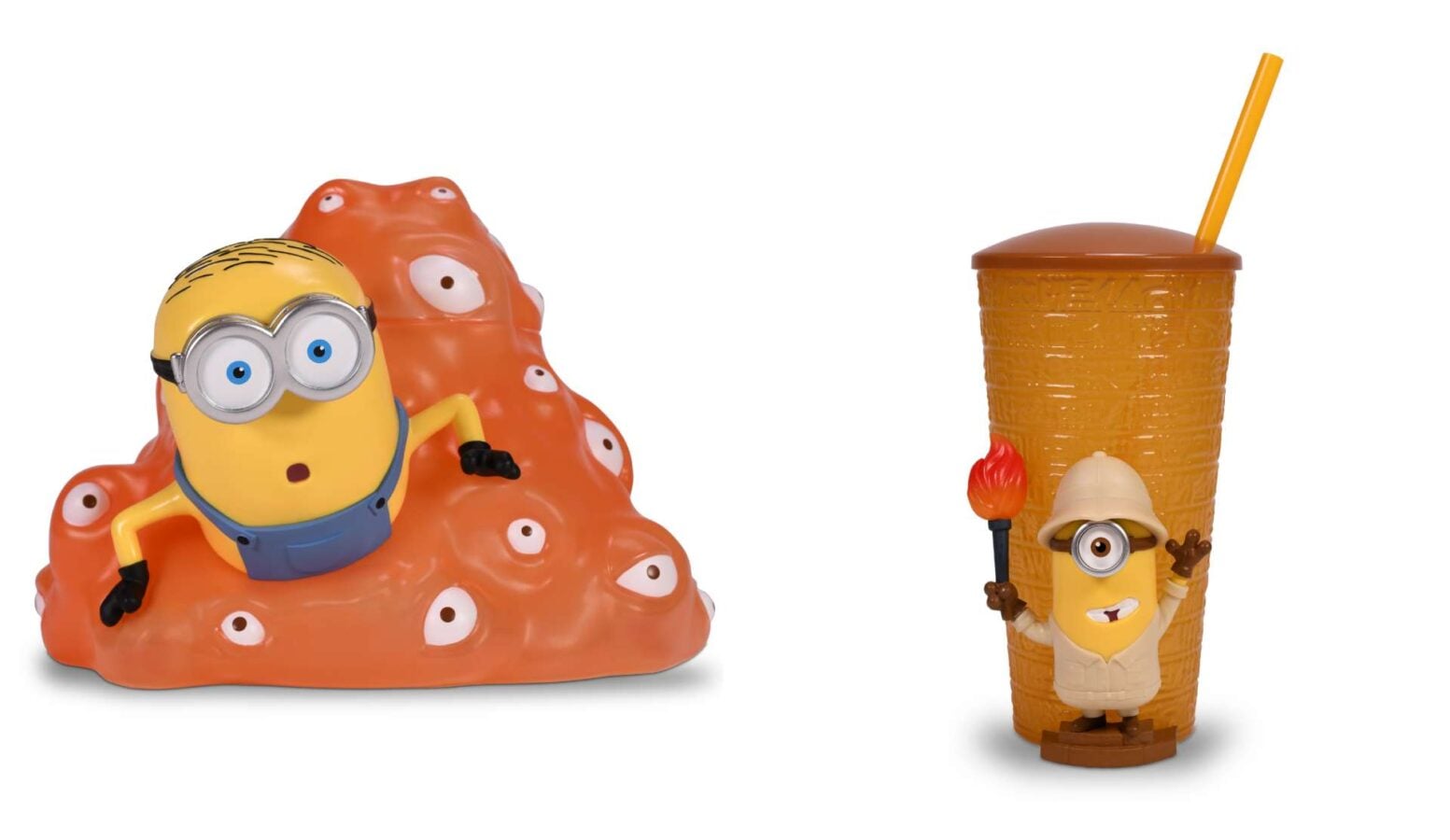 Two Minion figures sit with an orange bumpy mound and a tall orange cup with a straw in a playful toy scene
