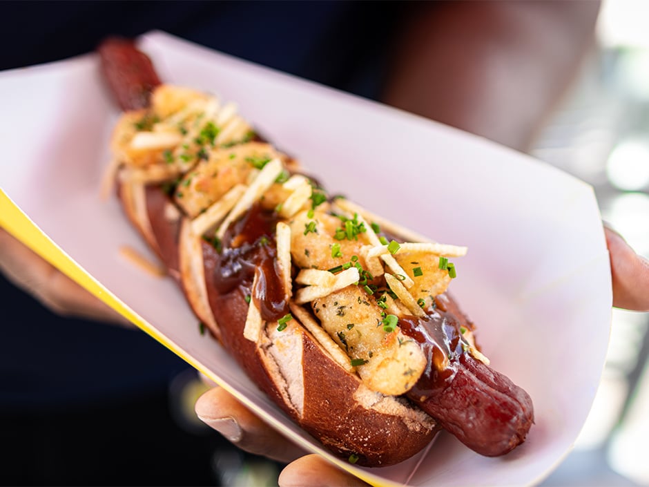 Hot dog in a bun topped with French fries fried chicken pieces barbecue sauce and chopped chives in a paper tray
