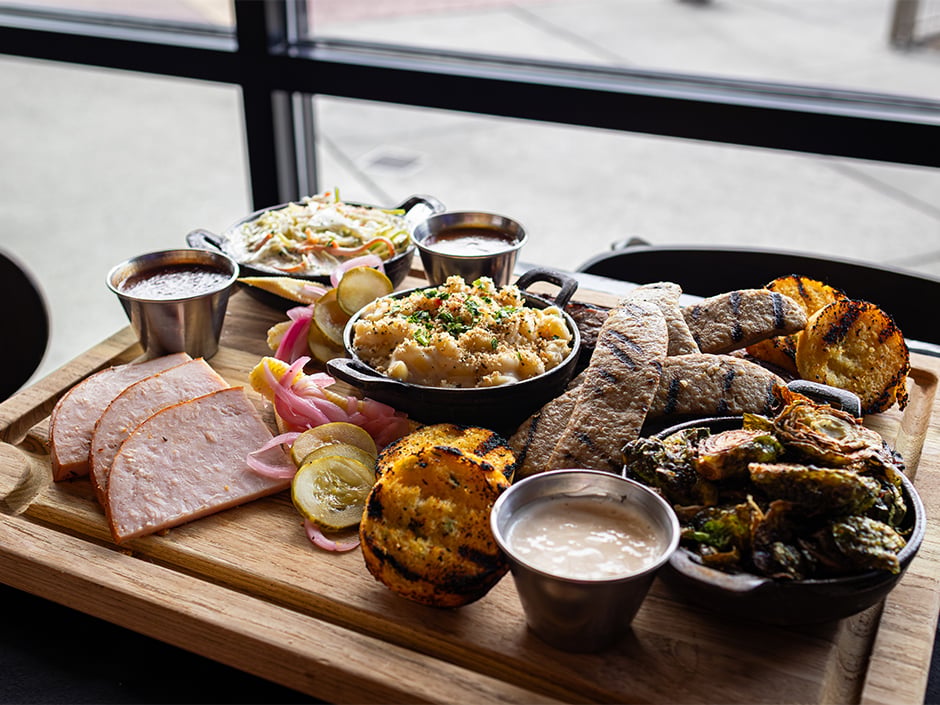 Charcuterie board with sliced ham pickles pickled onions roasted potatoes grilled vegetables and a skillet of macaroni and cheese with dipping sauces on the side