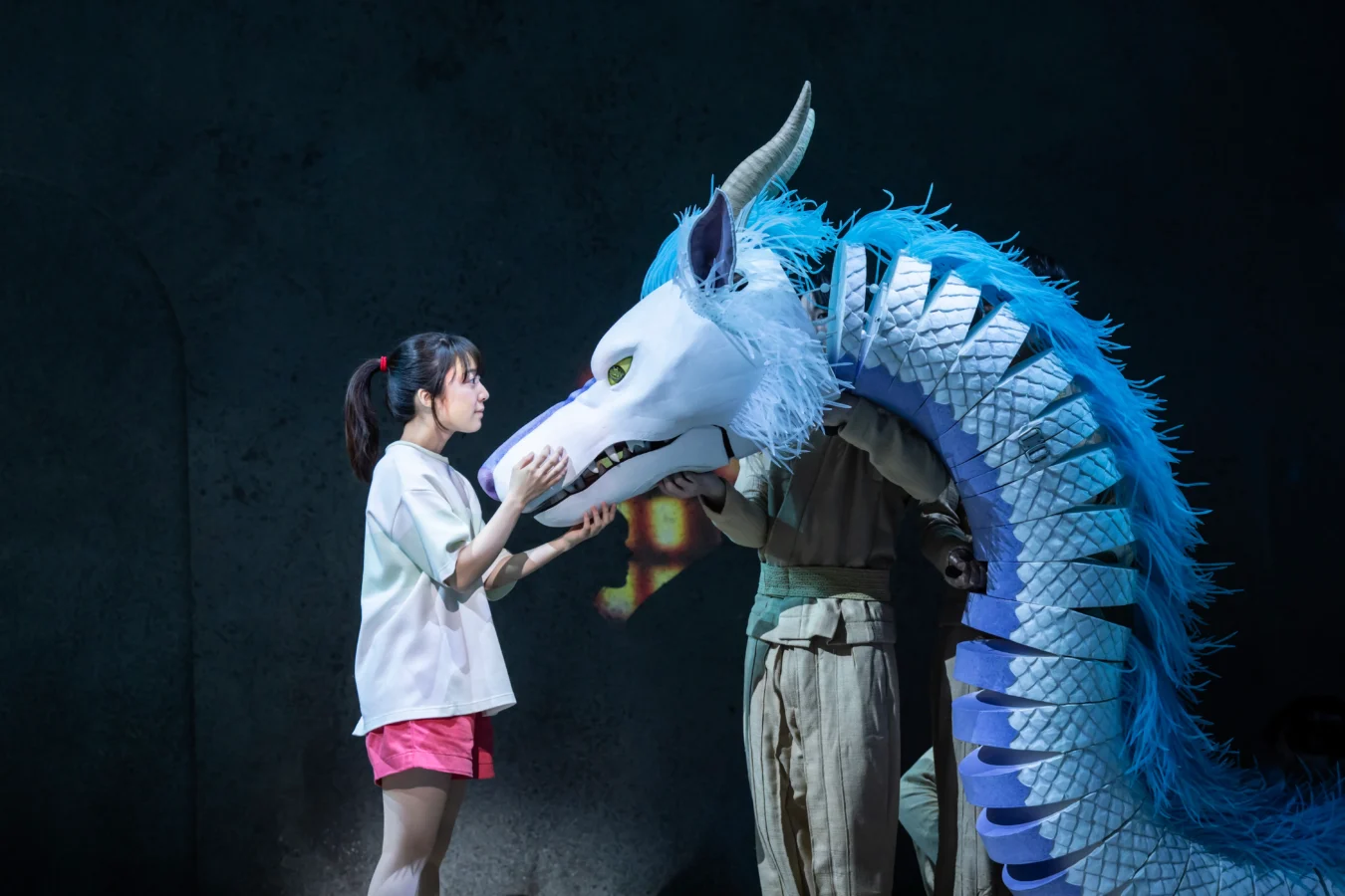 Girl in a white shirt and pink shorts cups the snout of a blue dragon costume against a dark stage backdrop sharing a quiet moment