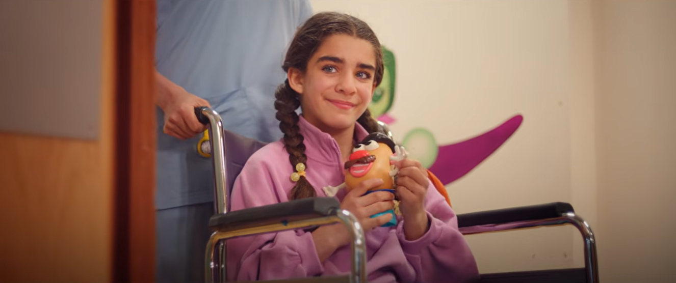 Screenshot 20260615 080329 Fantasy Land News Smiling girl in a pink hoodie sits in a wheelchair holding a colorful puppet as a caregiver pushes her down a hallway