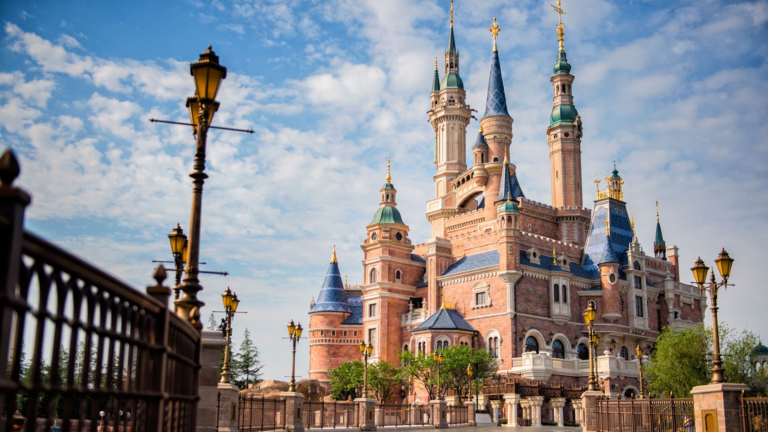 Fantasy castle with brick towers, blue roofs and gold spires, framed by a wrought-iron fence and decorative street lamps under a blue sky.