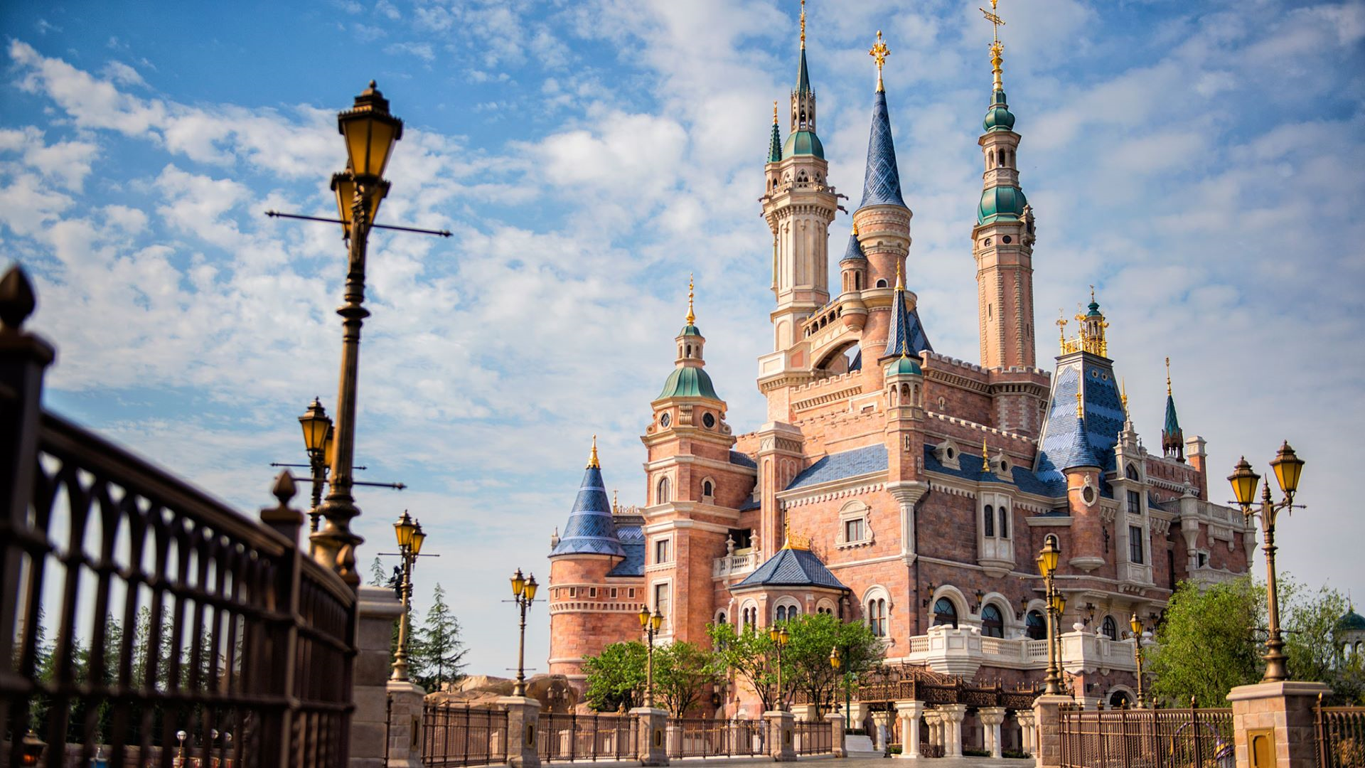 Fantasy castle with brick towers, blue roofs and gold spires, framed by a wrought-iron fence and decorative street lamps under a blue sky.