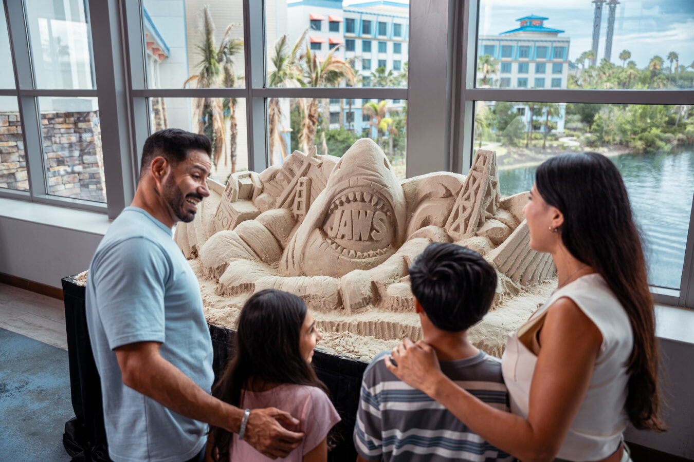 Group of people gathered around a large sand sculpture with the word JAWS carved in the center indoors by large windows overlooking city scenery