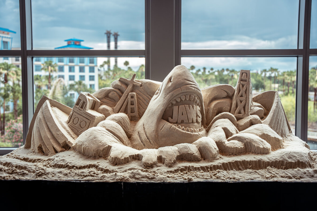 Sand sculpture on a display table inside a room with architectural carvings and the letters JAX carved in the sand seen through a window