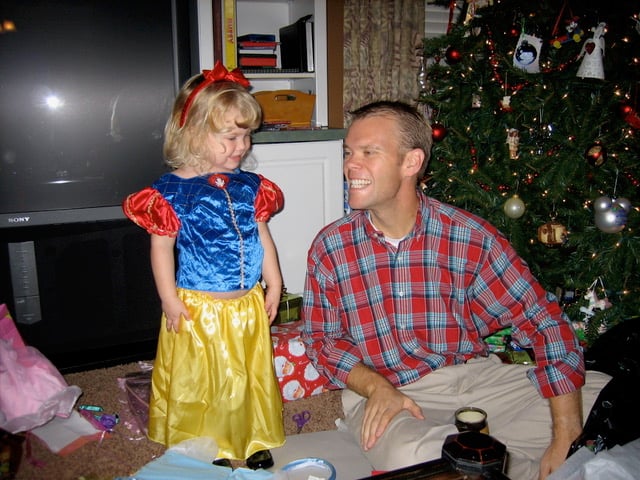 Little girl dressed as Snow White stands beside a smiling man in a plaid shirt near a Christmas tree