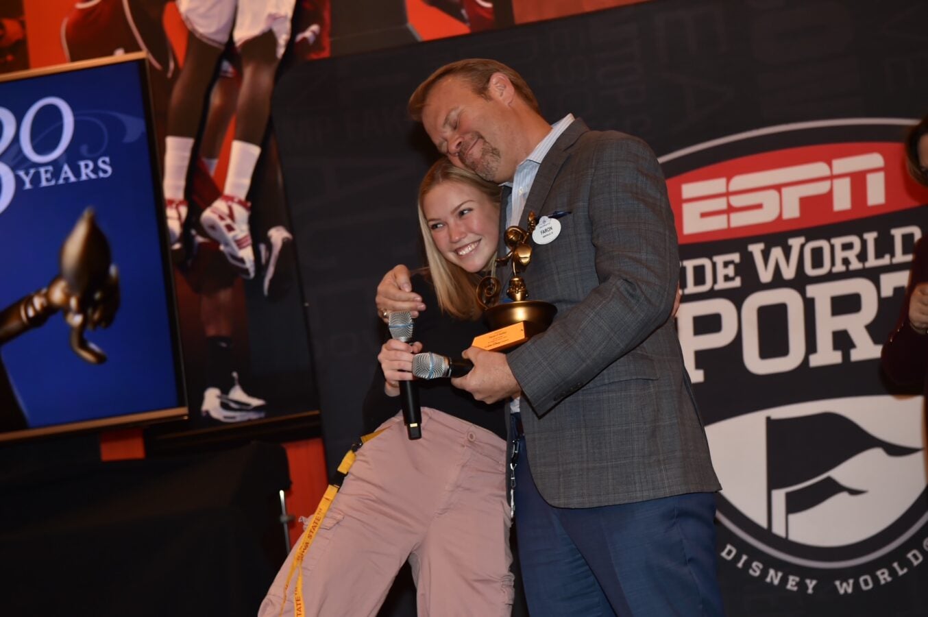 Man in a gray blazer hugs a smiling woman on a stage while holding a trophy with ESPN Wide World of Sports branding in the background