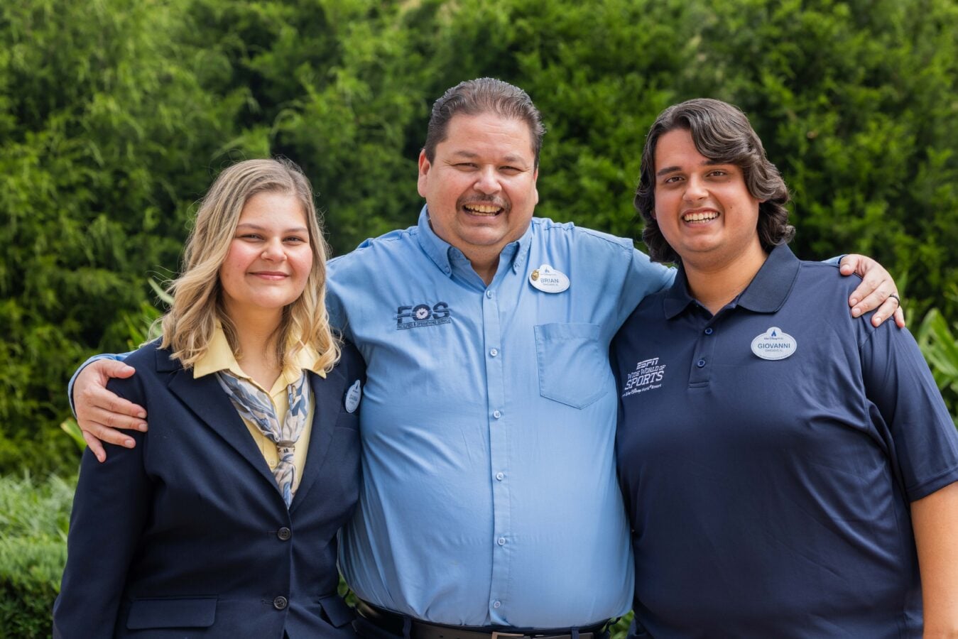 Three staff members with their arms around each other smiling outdoors in blue uniforms against a green hedge background
