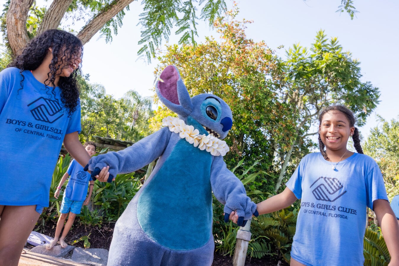 Kids in matching blue T shirts stand outdoors holding hands with a blue Stitch mascot wearing a flower lei