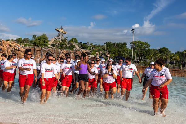 Group of volunteers running through knee deep water at a sunny pool area wearing white shirts and red shorts