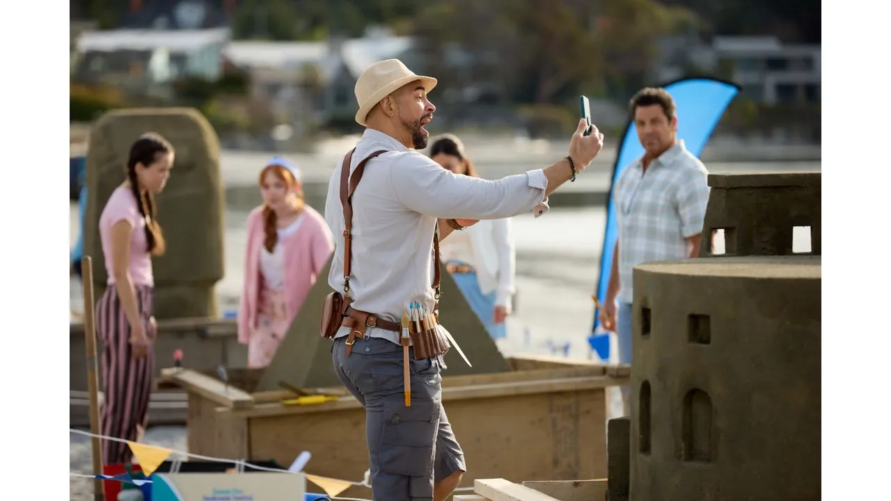 Man with a tool belt and hat takes a selfie at an outdoor beachside construction site coworkers in the background