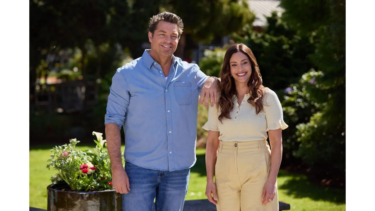 Smiling man in a blue shirt and jeans stands with a smiling woman in a cream top and beige pants in a sunny garden