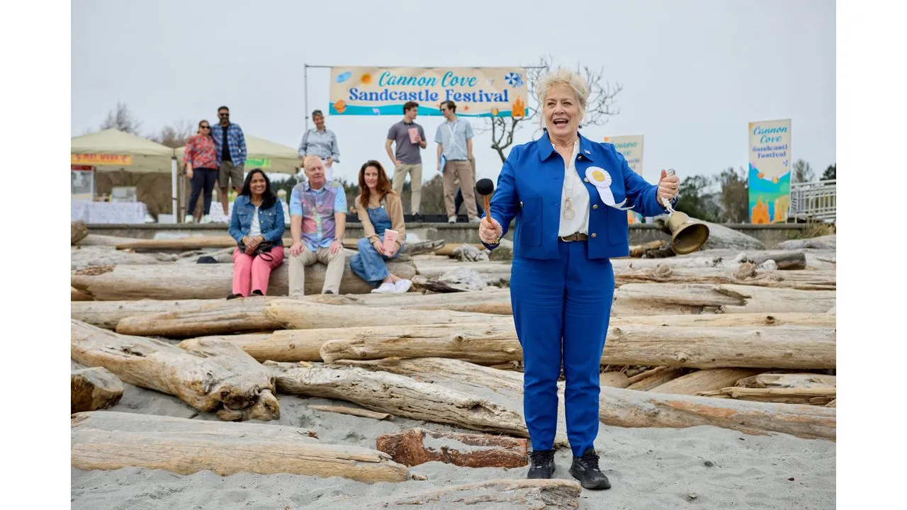 Woman in a blue suit addresses a crowd with a microphone on a driftwood stage at Cannon Cove Sandcastle Festival