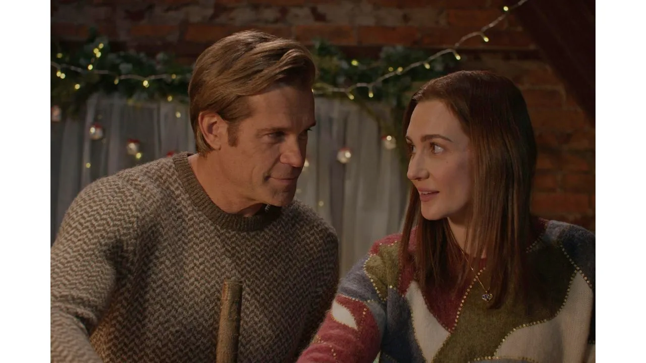 A man and a woman sit together smiling and talking in a cozy festive room with string lights in the background