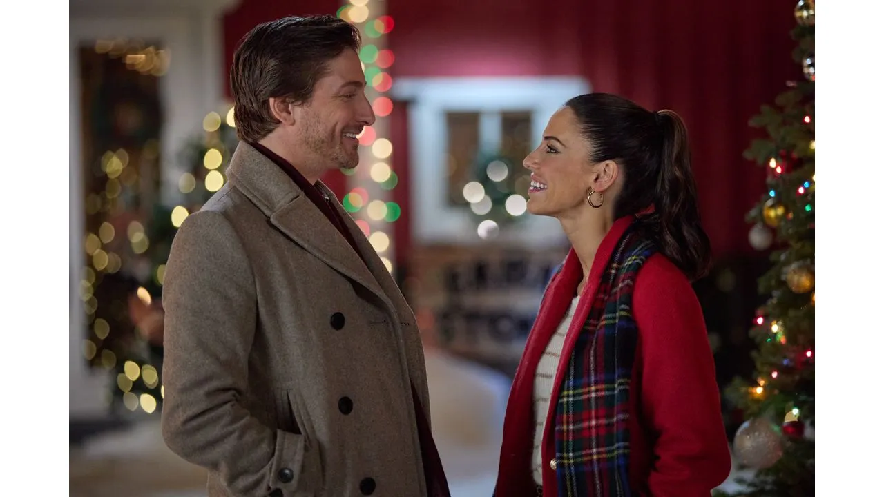 A man and a woman smile at each other indoors wearing warm coats with festive lights and a Christmas tree in the background