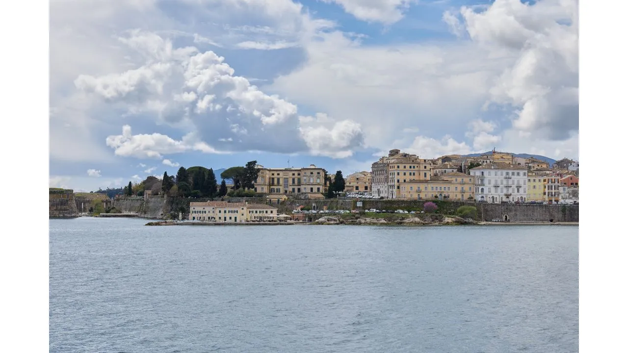 greek5 Fantasy Land News Coastal town along a calm bay with pastel buildings and a stone seawall under a cloudy sky seen across the water
