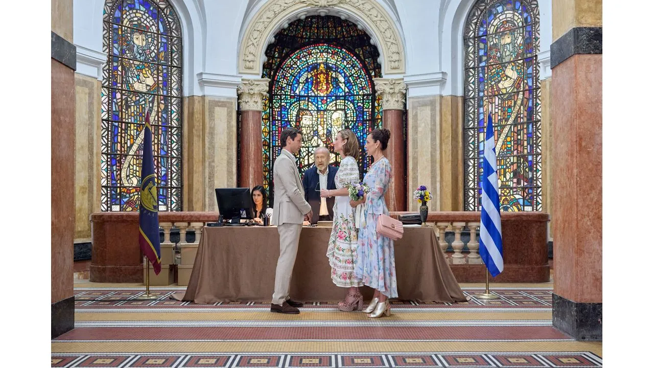 greek7 Fantasy Land News Man in a light suit shakes hands with a woman in a floral dress during a formal ceremony inside a grand stained glass hall an official and a staffer are at a table behind them