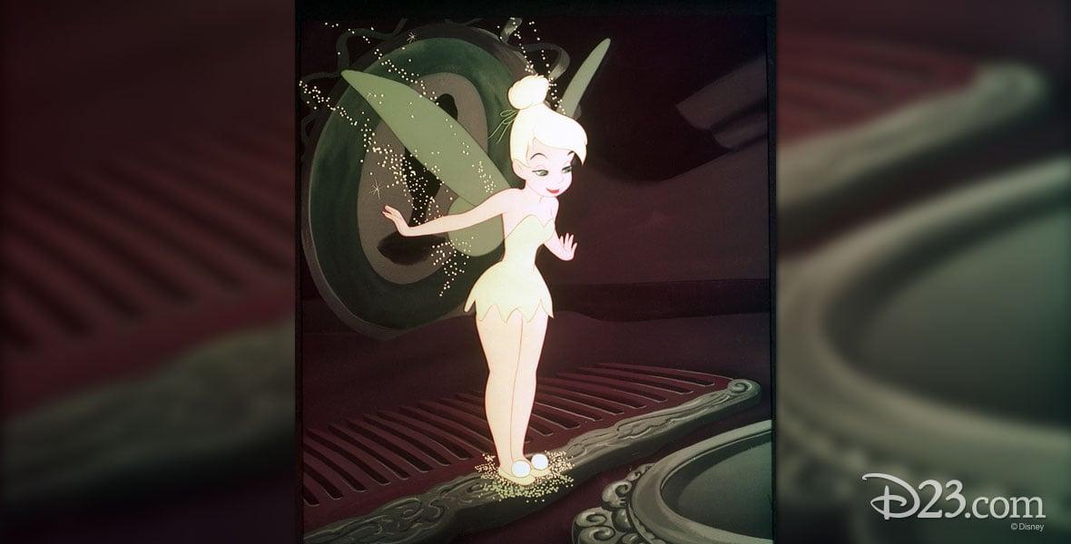 Tinker Bell in a yellow dress stands on a decorative green surface sprinkling pixie dust