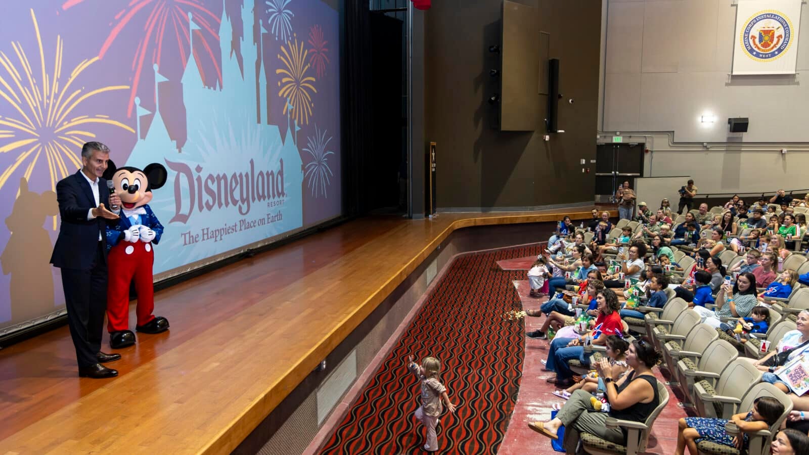 Man and Mickey Mouse on stage at a Disneyland event addressing a seated audience