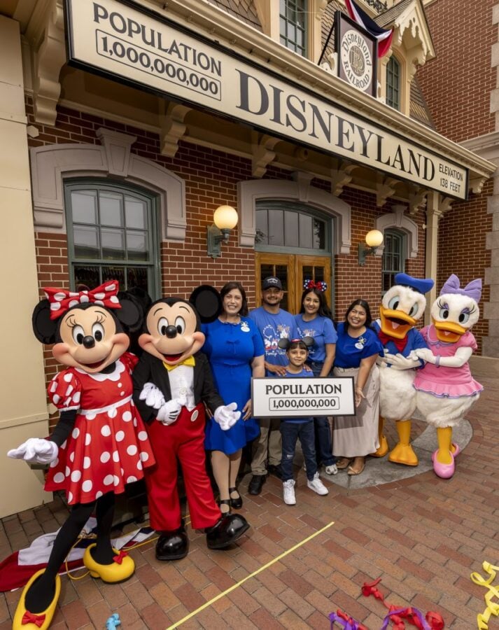 Group of people and Disney characters pose outside Disneyland holding a sign that reads Population 1000000000