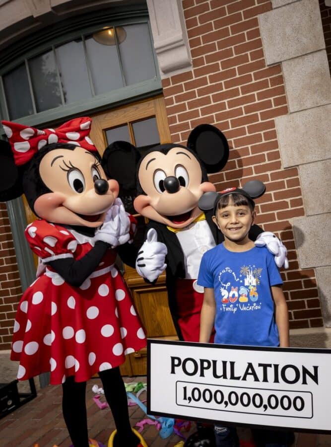 Boy stands with Mickey and Minnie Mouse outside a brick building smiling beside a sign that reads Population 1000000000