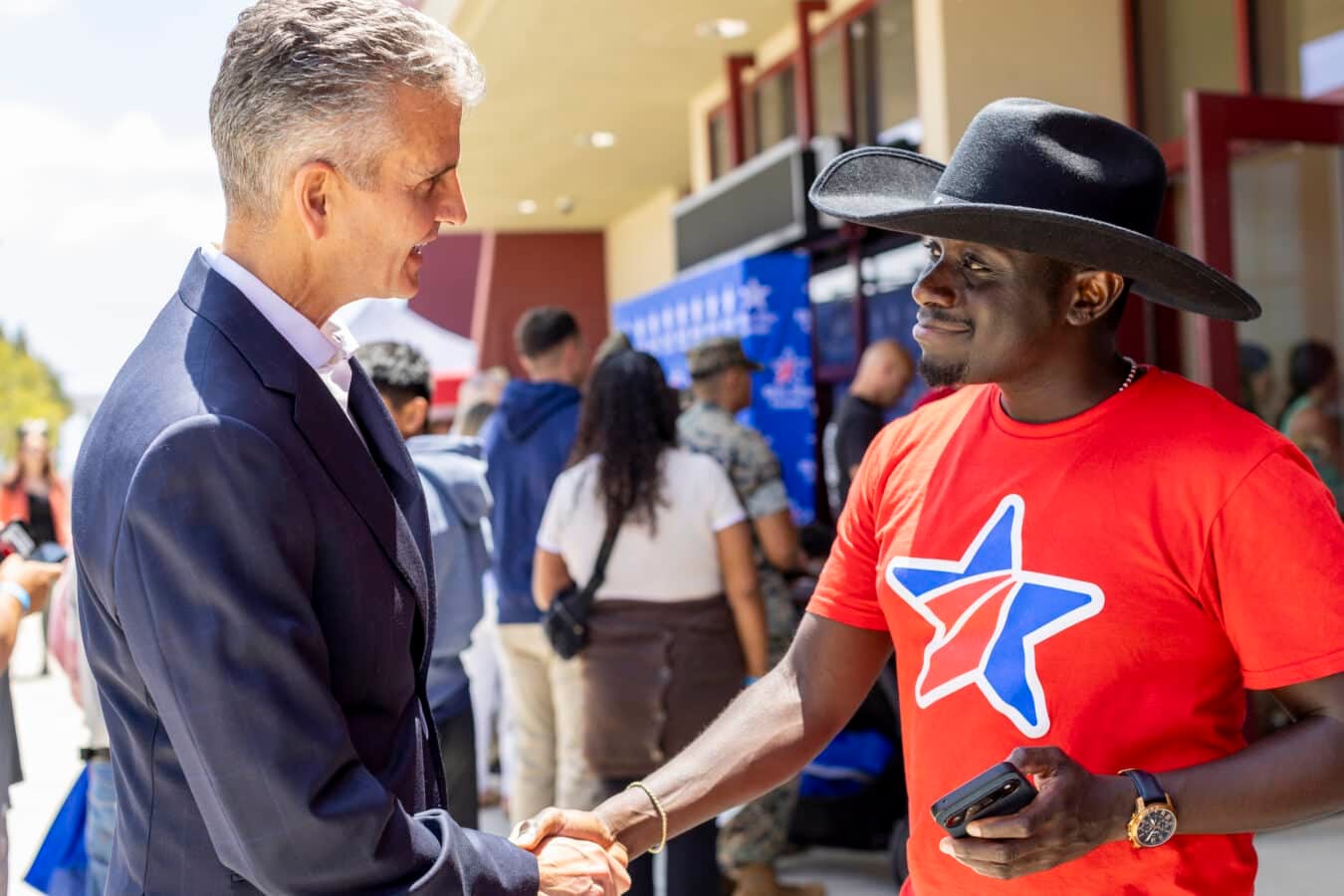 Two men shake hands at an outdoor event one in a navy suit the other in a red shirt with a star graphic and a wide brim black hat