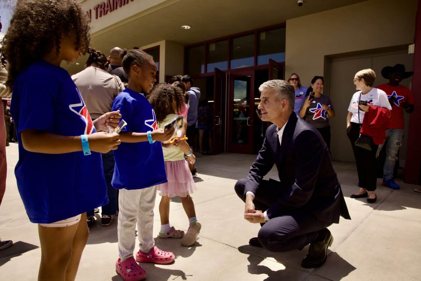 Man in a suit kneels and speaks to children lined up outside a building as volunteers hand out items