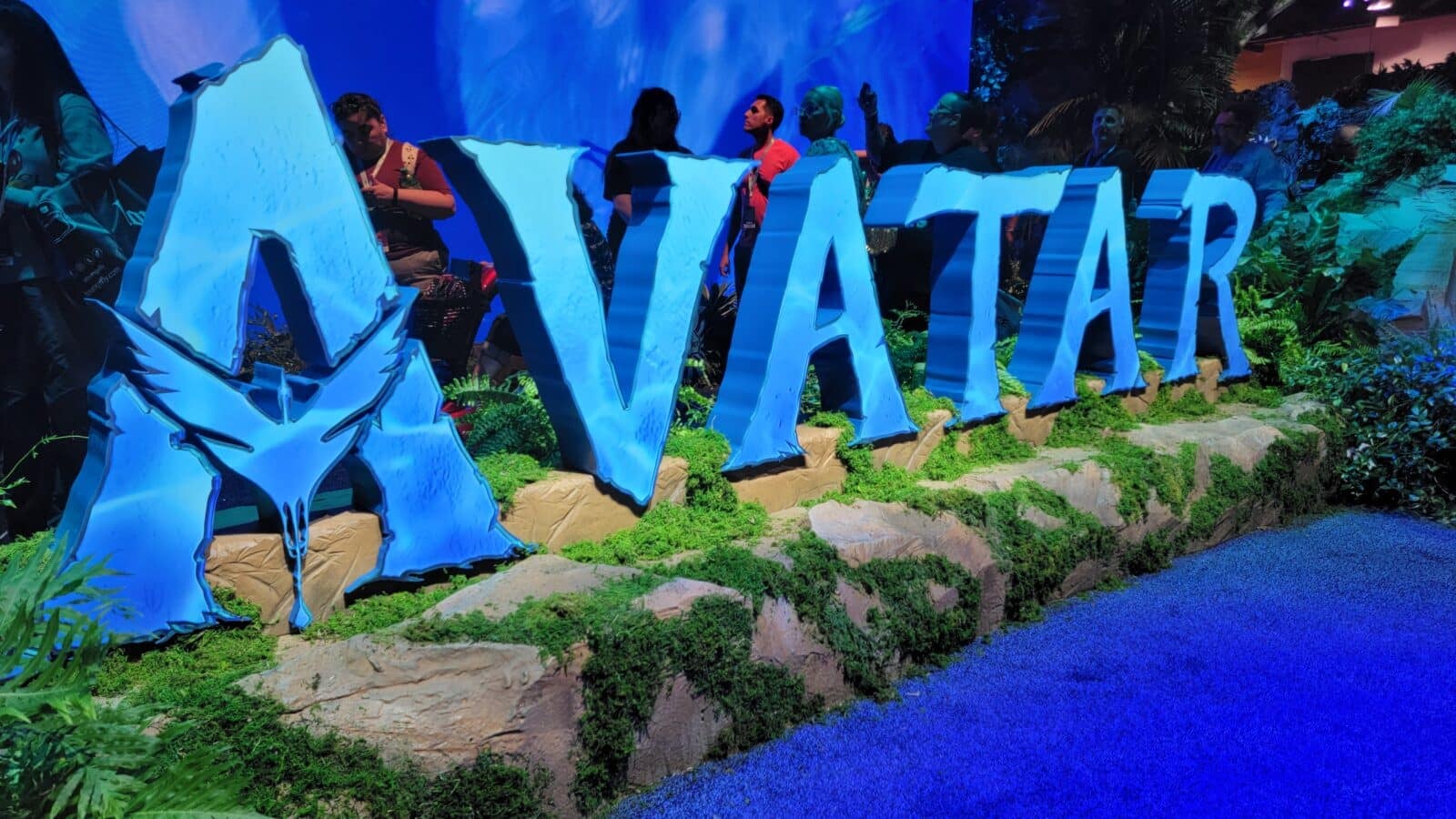 Oversized blue letters spell AVATAR at a nighttime exhibit with visitors standing behind the sign among plants