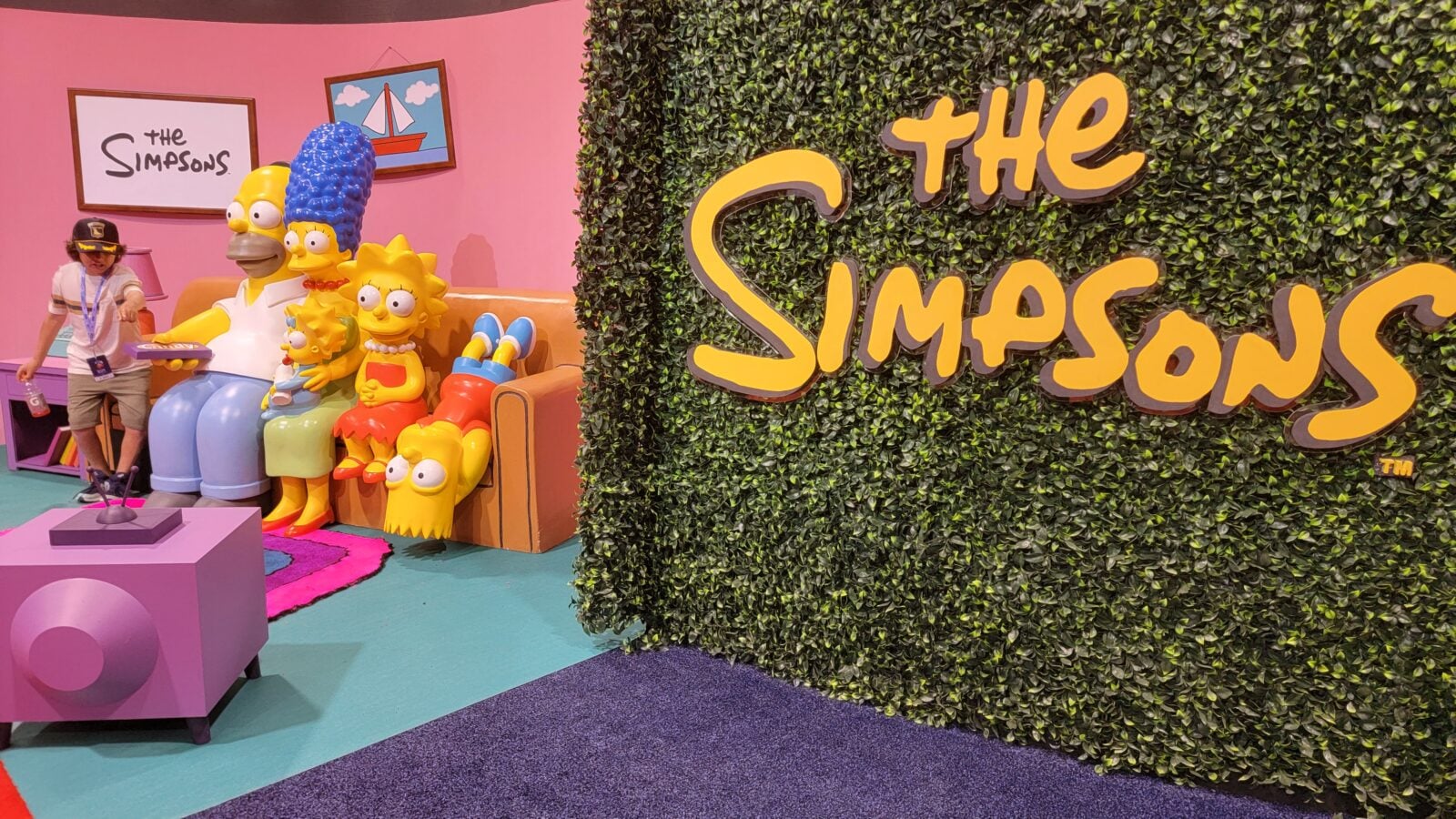 Life size Simpsons family figures Homer Marge Bart Lisa Maggie on a yellow couch in a pink room with a large The Simpsons logo on a leafy green wall