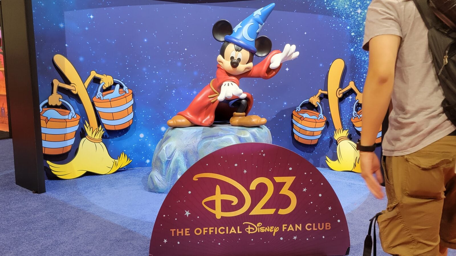 Mickey Mouse in a blue wizard hat and red robe poses on a rocky pedestal against a starry blue background with a Disney Fan Club sign in front