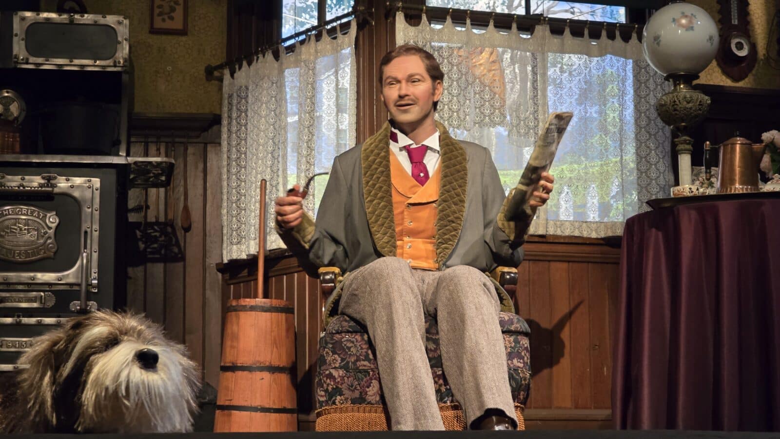 Actor in Victorian style suit seated on a stage holding a pipe and newspaper with a dog visible in the foreground