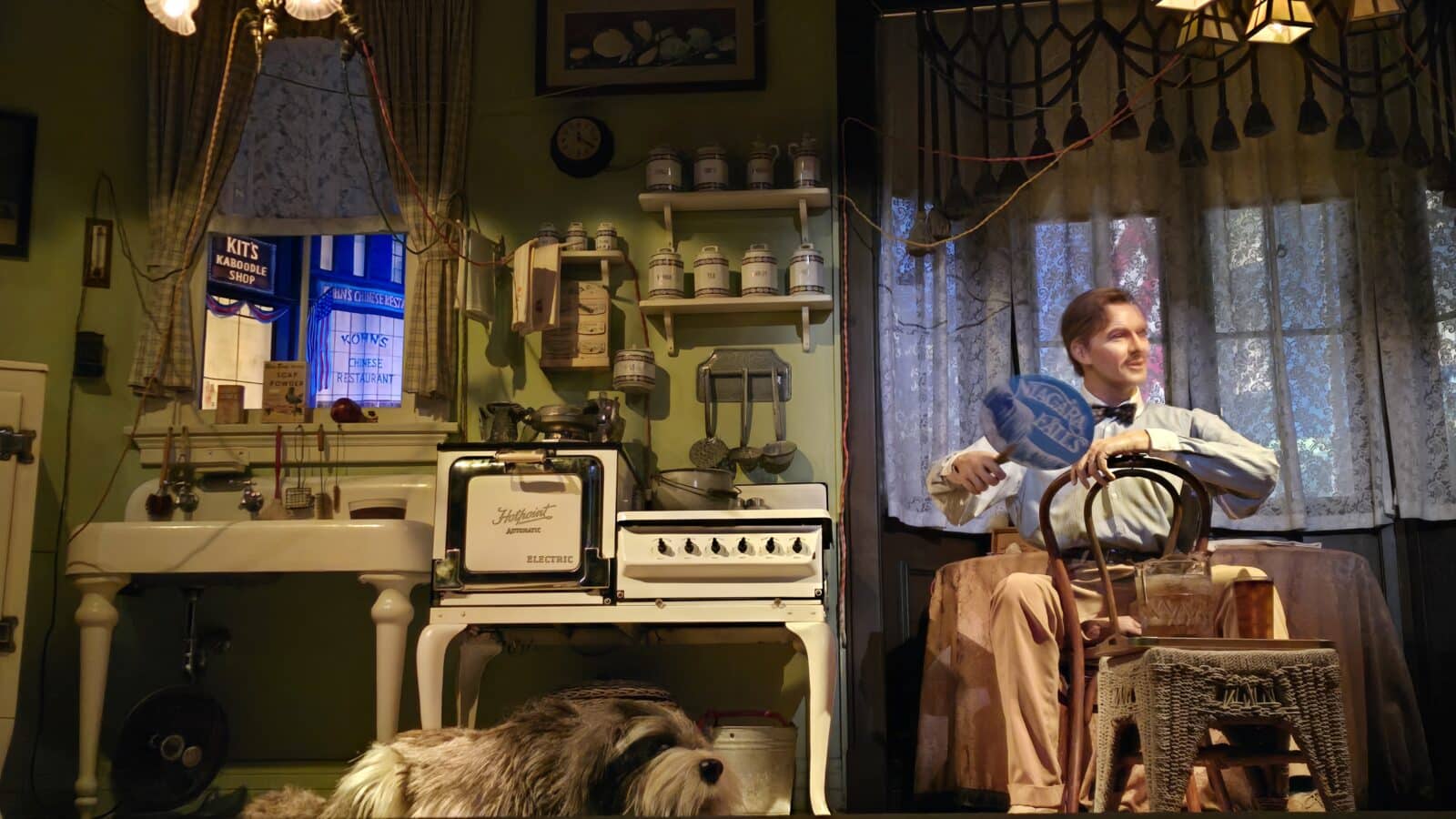 A vintage kitchen scene with a man in a bow tie seated on a chair holding a blue fan while a dog rests in the foreground near a retro stove and shelves of jars
