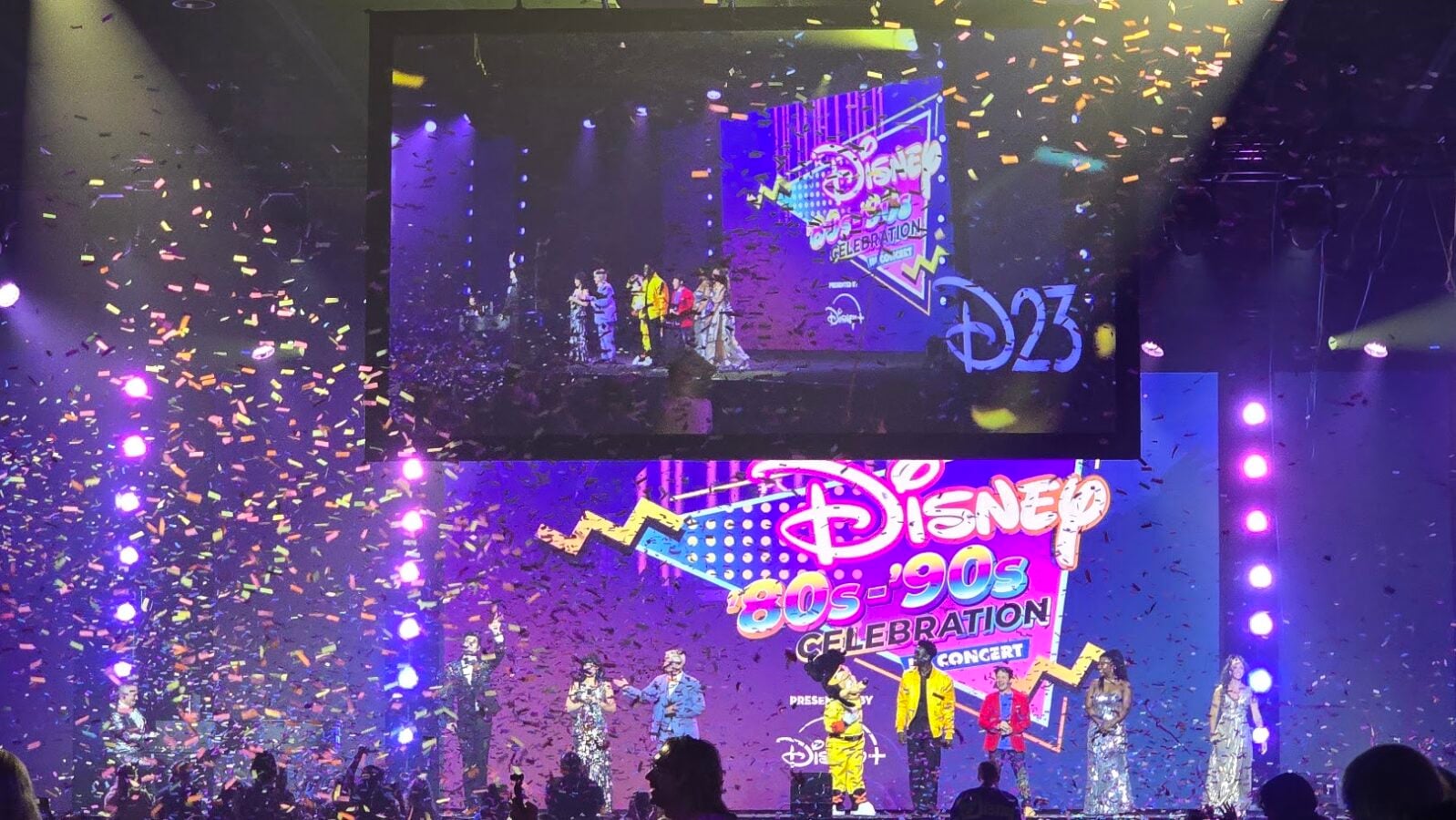 Stage at a Disney 80s90s Celebration concert with performers neon backdrop and colorful confetti raining down