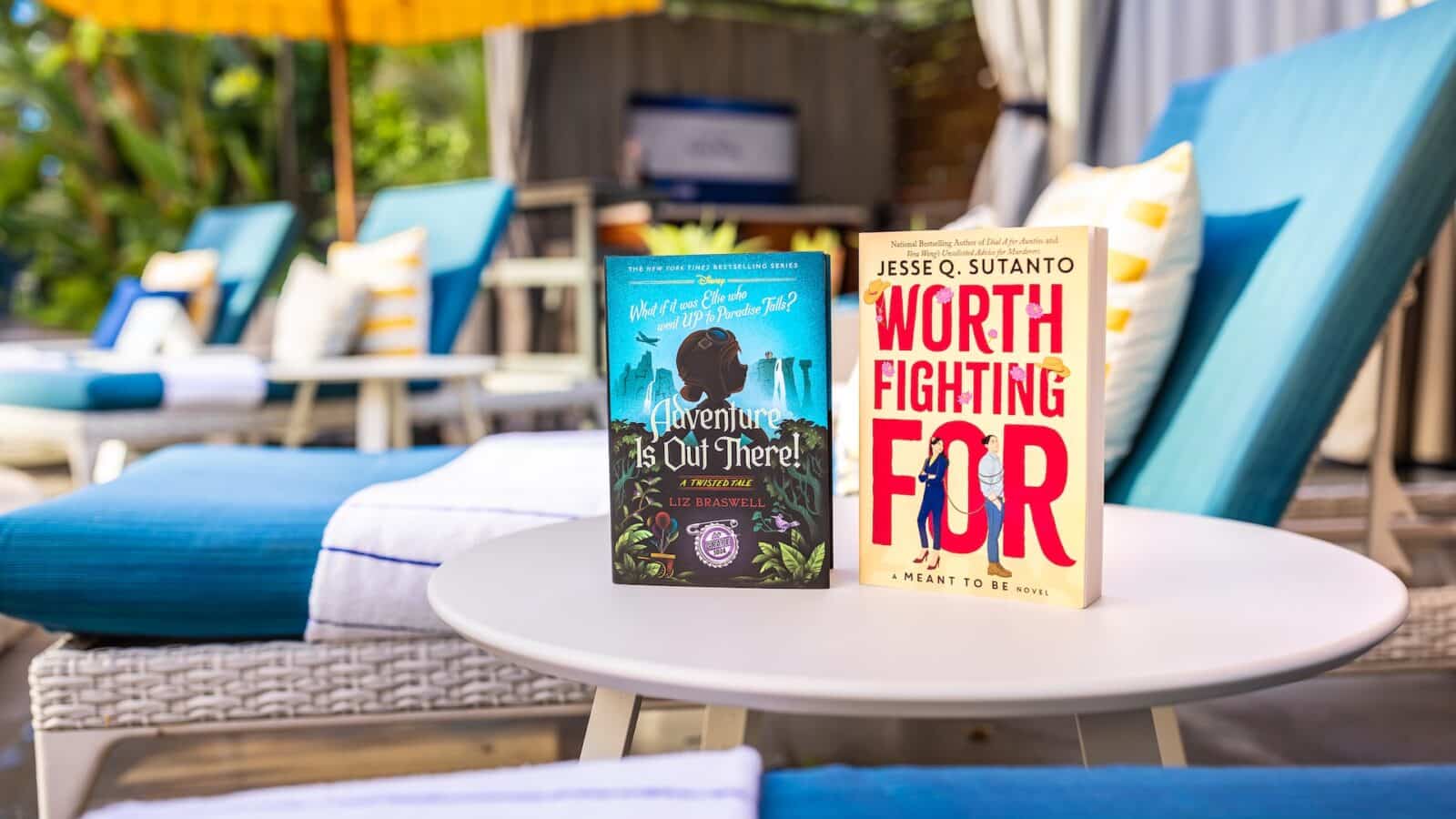 Two paperback books on a white round table beside blue poolside lounge chairs in bright sunny setting