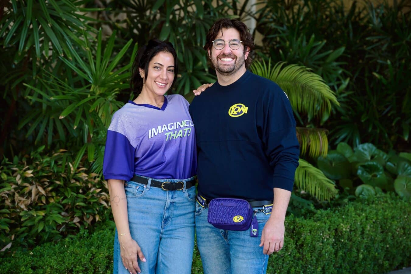 Two smiling adults stand close together in a lush garden arms around each other wearing casual purple and navy shirts