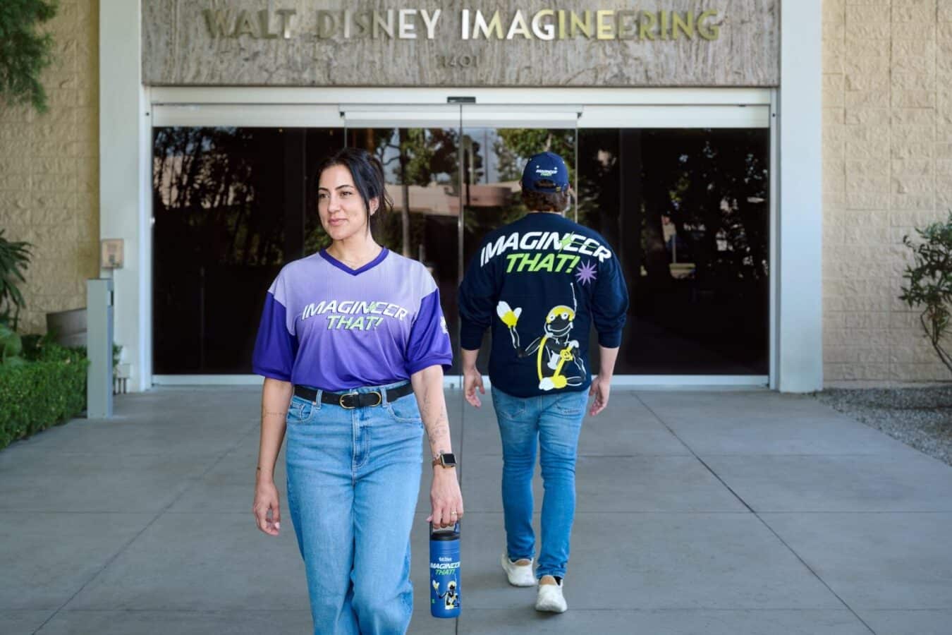 Two people wearing Imagineer That shirts walk outside a building labeled Walt Disney Imagineering the woman in front carries a blue water bottle
