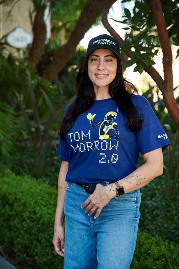 Smiling woman in a blue Tshirt with a robot graphic stands outdoors among green plants wearing a black cap that reads IMAGINE THAT