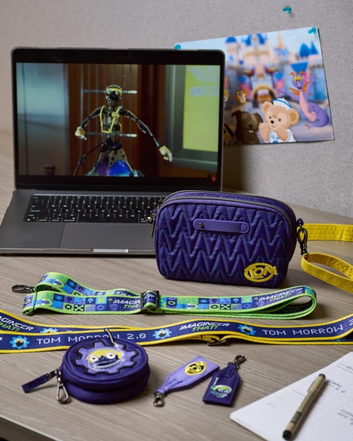 Laptop screen shows a toy robot figure on display with a purple quilted bag and colorful straps on a desk surface