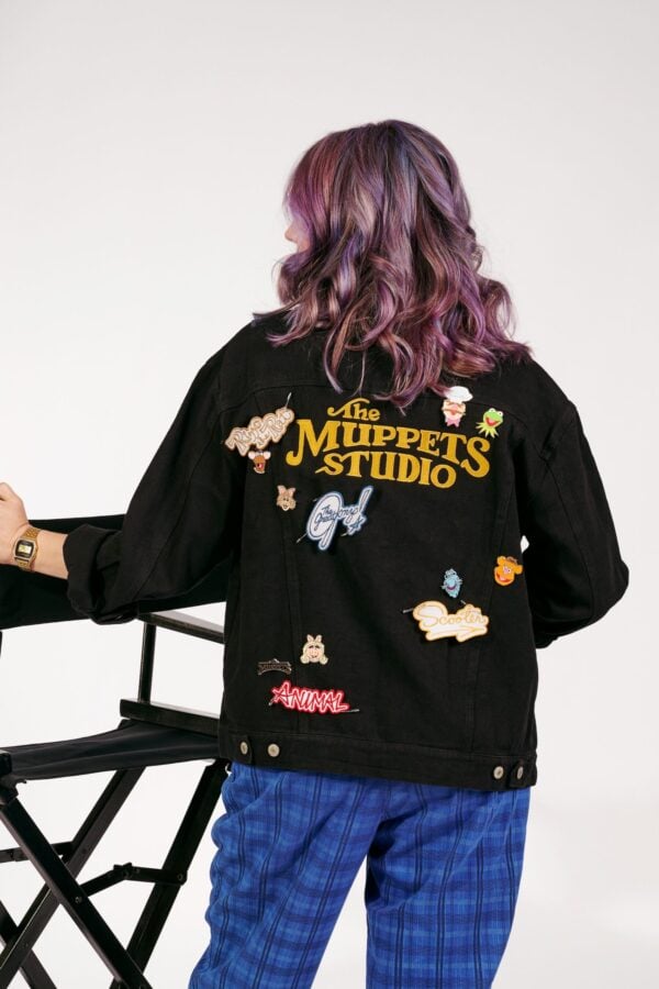 Person with shoulder length purple dyed hair wearing a black jacket covered in Muppets Studio patches seated on a directors chair seen from the back