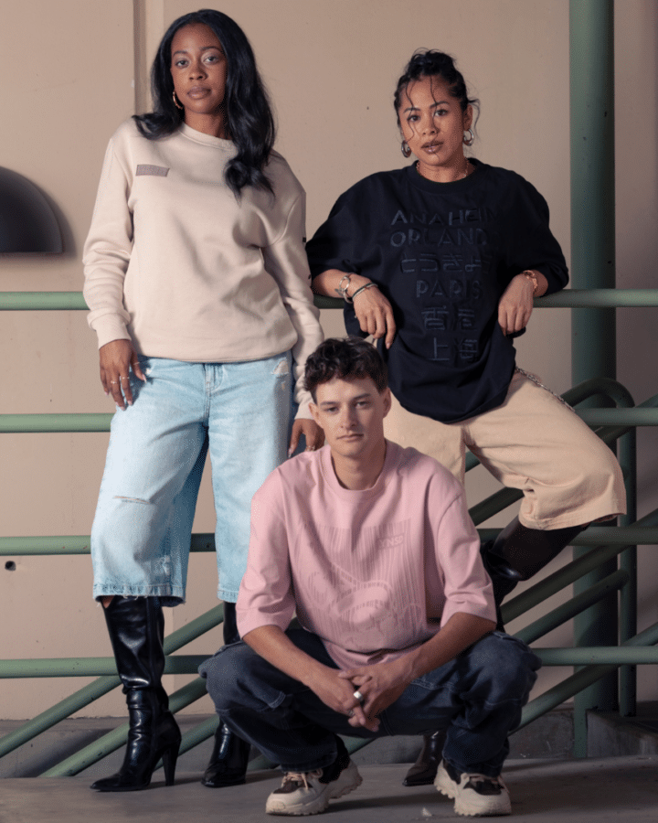 Group portrait of three friends posing on a green railing two standing one in a beige sweatshirt and light jeans with knee high boots one in a black oversized shirt and tan pants and a third person kneeling in a pink oversized shirt and dark jeans