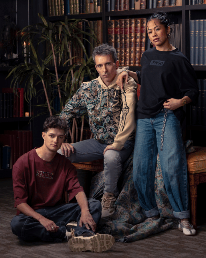 Three people pose in a cozy library setting with tall bookshelves one sits on the floor another sits on a bench and a third stands beside them with a hand on the bench