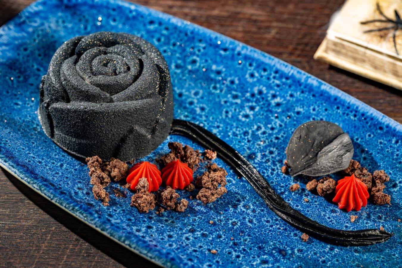 Black sugar dusted rose shaped dessert on a blue speckled plate with red piped frosting accents and chocolate crumbs