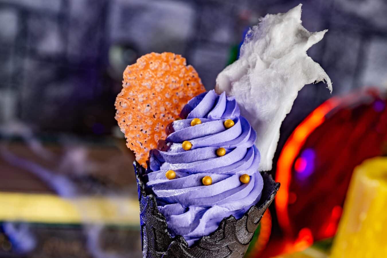 Close up of a dessert cone with purple frosting orange crispy cookie and white cotton candy garnish