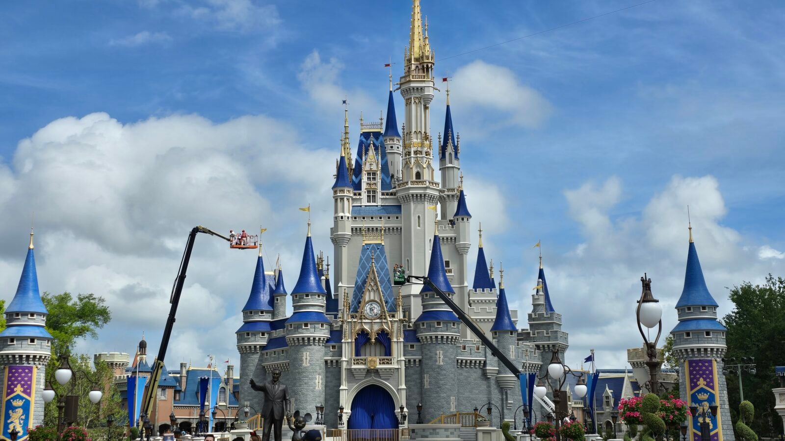 Front view of a large fantasy castle with blue tiled turrets and gold accents against a blue sky with clouds A Disney like castle with tall spires and flags rises above a courtyard scene Grand castle featuring blue roofs intricate arches and tall golden spires at a theme park