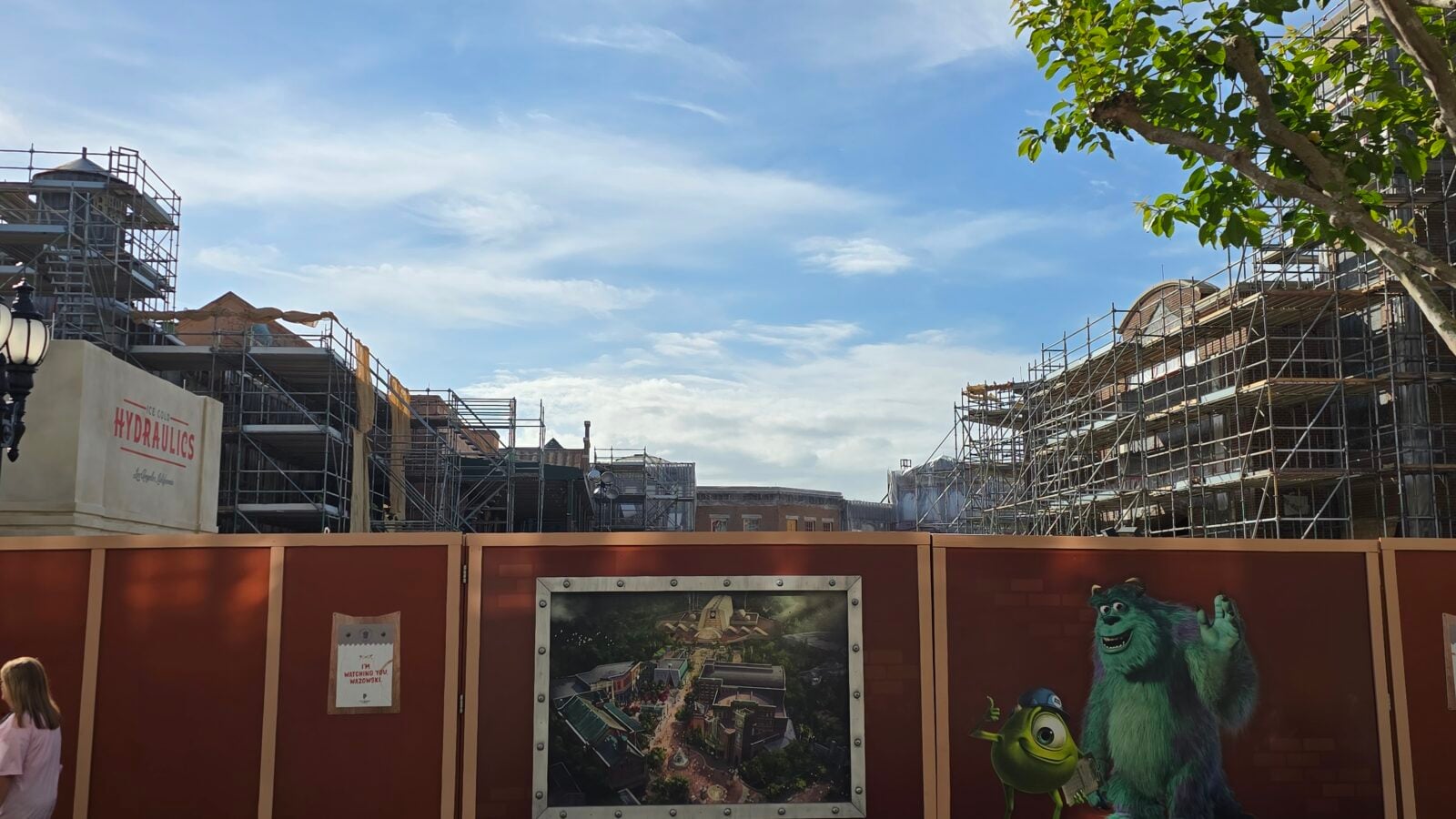Active construction site with metal scaffolding on both sides and a clear blue sky above mural of Mike and Sully from Monsters Inc on the fence