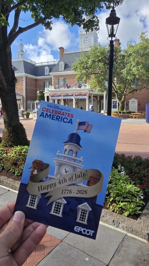 Hand holding a Disney World Celebrates America poster with a clock tower building in a sunny park backdrop