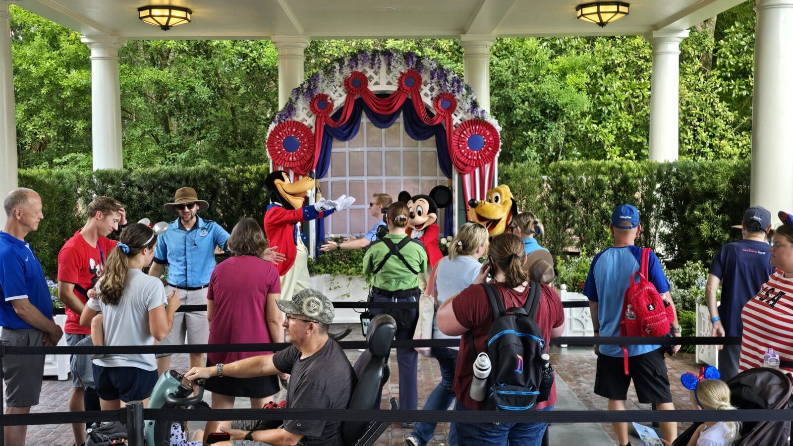 Disney characters Mickey Mouse and Goofy greet a crowd on a patriotic flower decorated stage under a gazebo