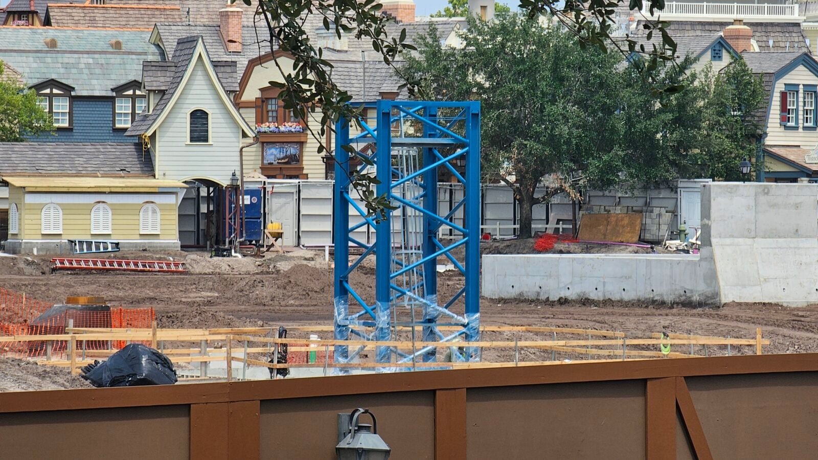 Construction site with blue metal scaffolding in the foreground and a row of pastel houses in the background Dirt ground and orange safety fencing visible