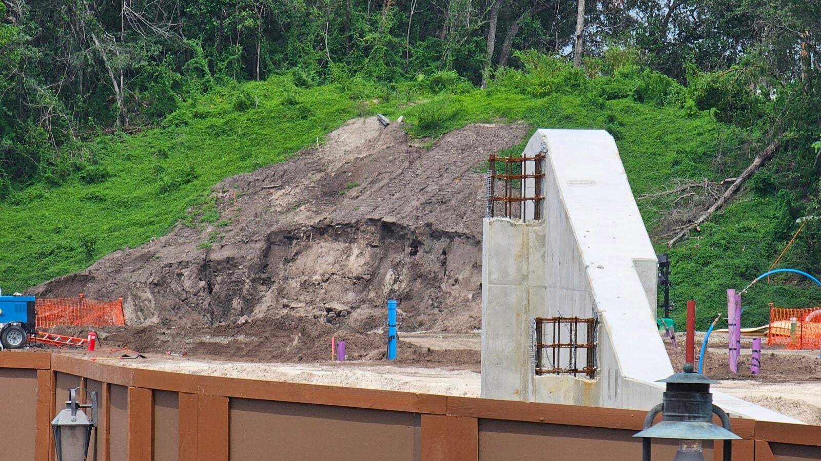 White concrete ramp under construction beside a dirt embankment and green hill with orange safety fencing
