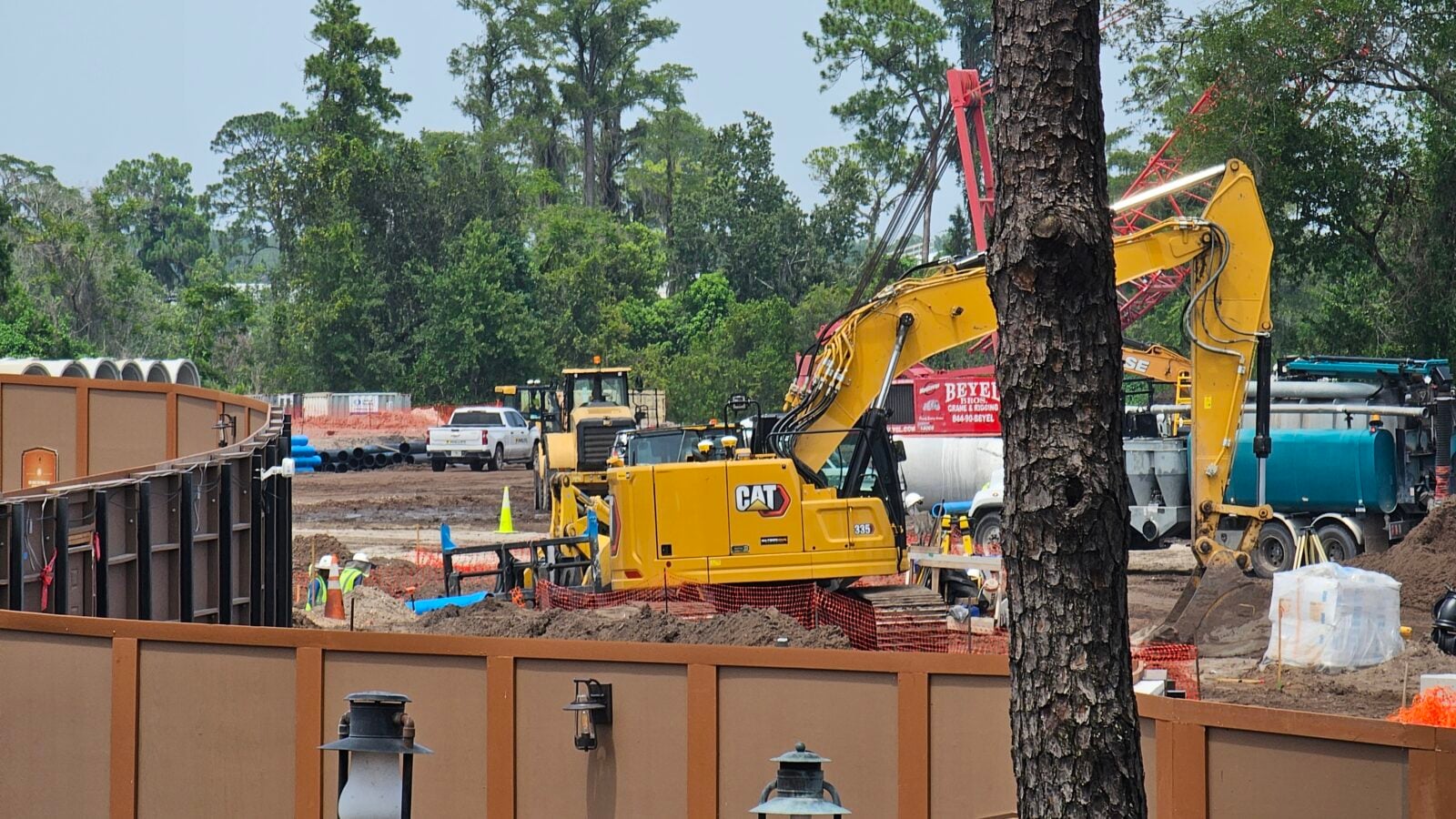 Construction site with yellow excavator and orange safety fencing amid dirt and trees in the background
